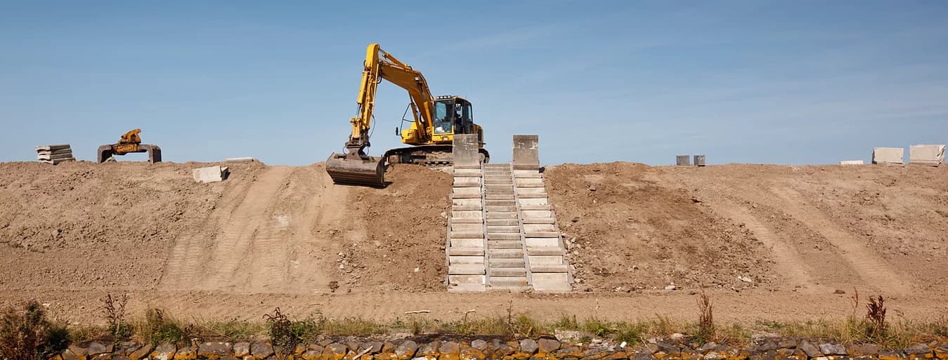 A yellow excavator atop a dirt embankment beside concrete-block steps under a clear blue sky.