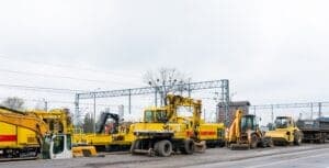 Excavators and bulldozers parked along a road near railway tracks under an overcast sky.