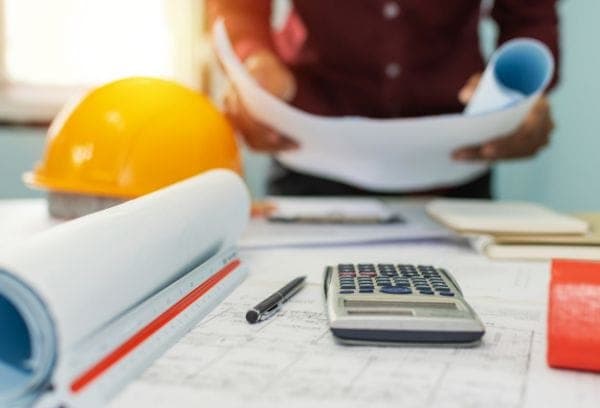 A person examines rolled blueprints on a desk with a calculator, pen, and orange hard hat nearby, suggesting an office or construction planning environment.