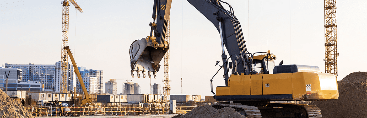 An excavator lifts dirt on a construction site, surrounded by cranes and a city skyline in the background, under a clear sky.