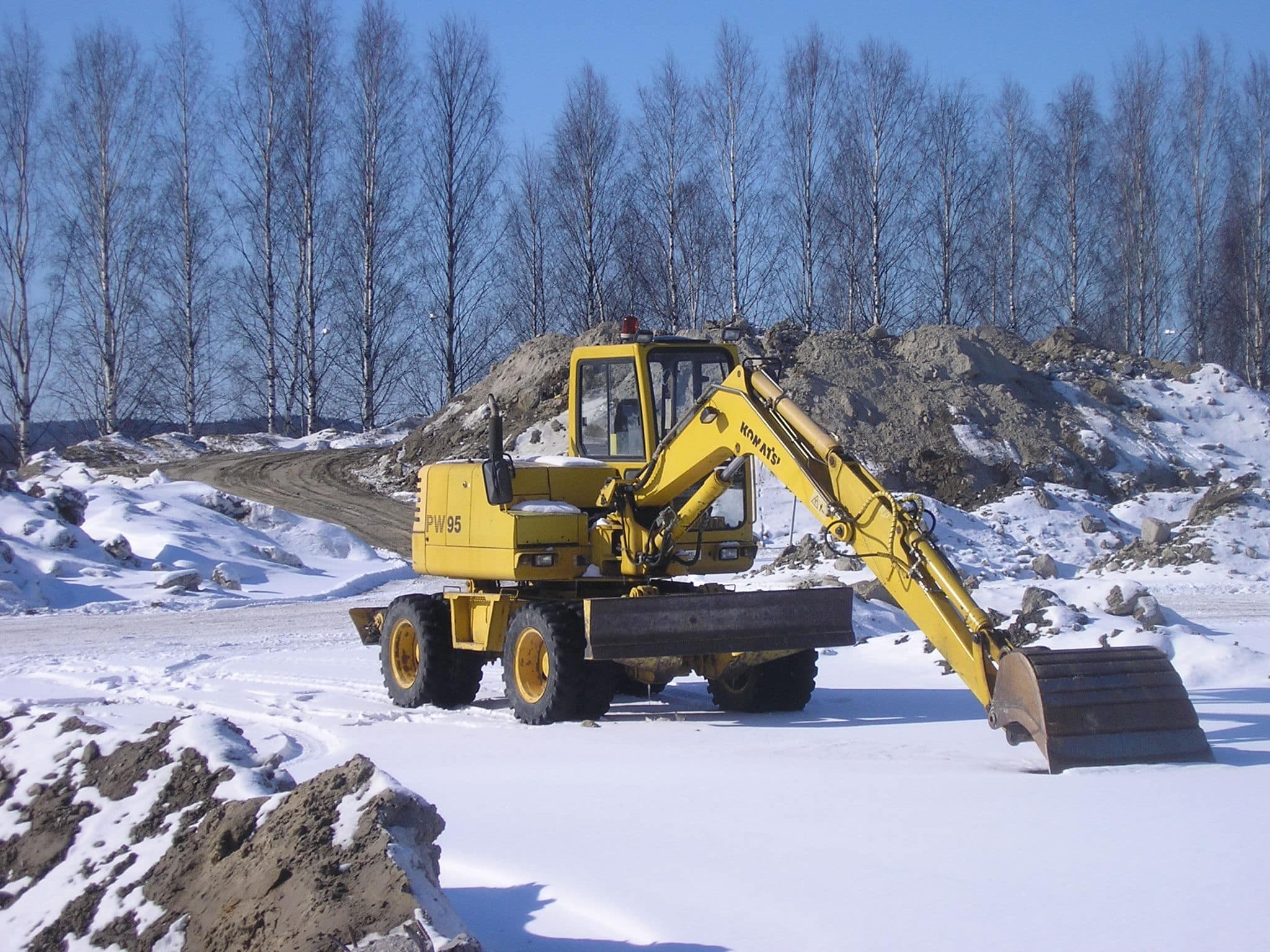 A yellow excavator on snow-covered ground with mounds of snow and dirt and leafless trees behind.
