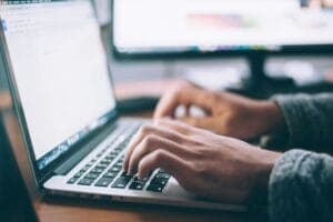 Hands typing on a laptop keyboard open to a text document, with blurred computer screens behind.