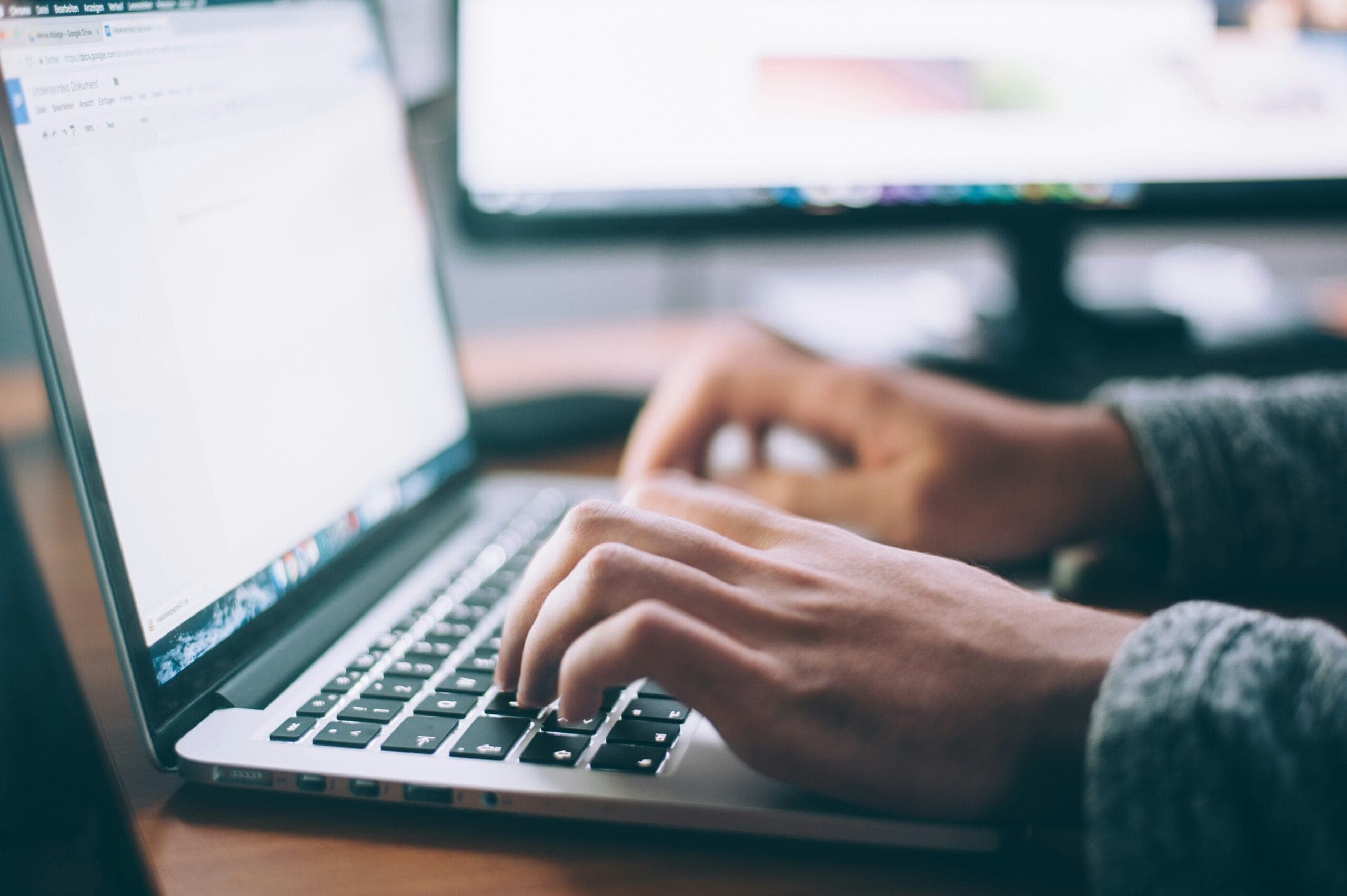 Hands typing on a laptop keyboard open to a text document, with blurred computer screens behind.