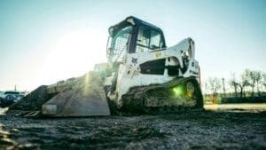 A skid steer loader on a construction site, surrounded by dirt with a sparse line of trees behind.