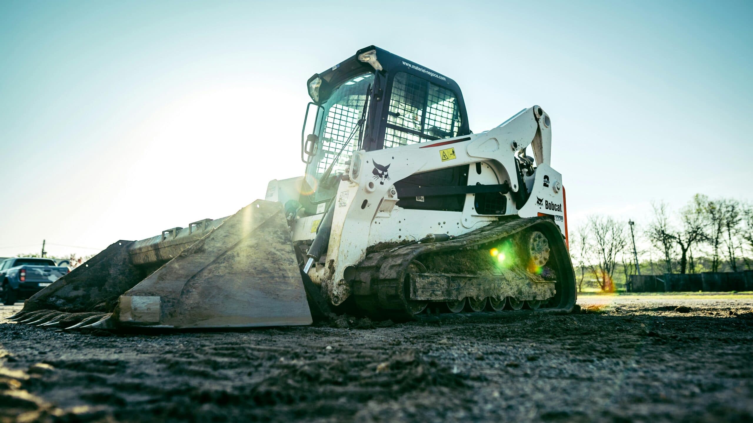A skid steer loader rests on a construction site, bathed in sunlight, surrounded by dirt and a sparse line of trees in the background.