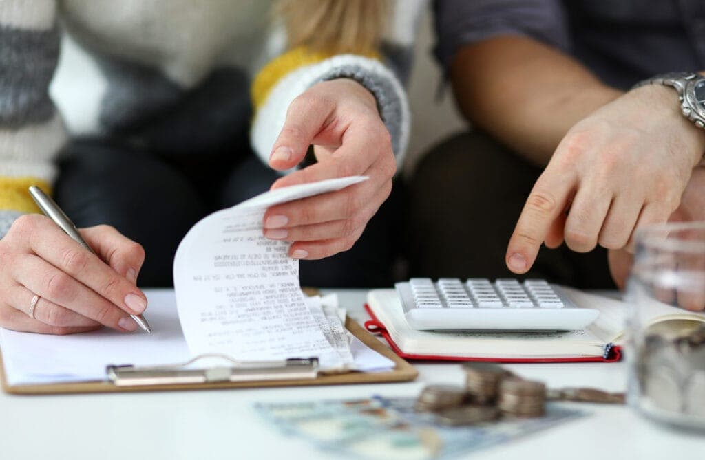 Two people sit at a table, one writing on a clipboard and the other pointing at a calculator. Coins and currency are spread around, suggesting financial planning.