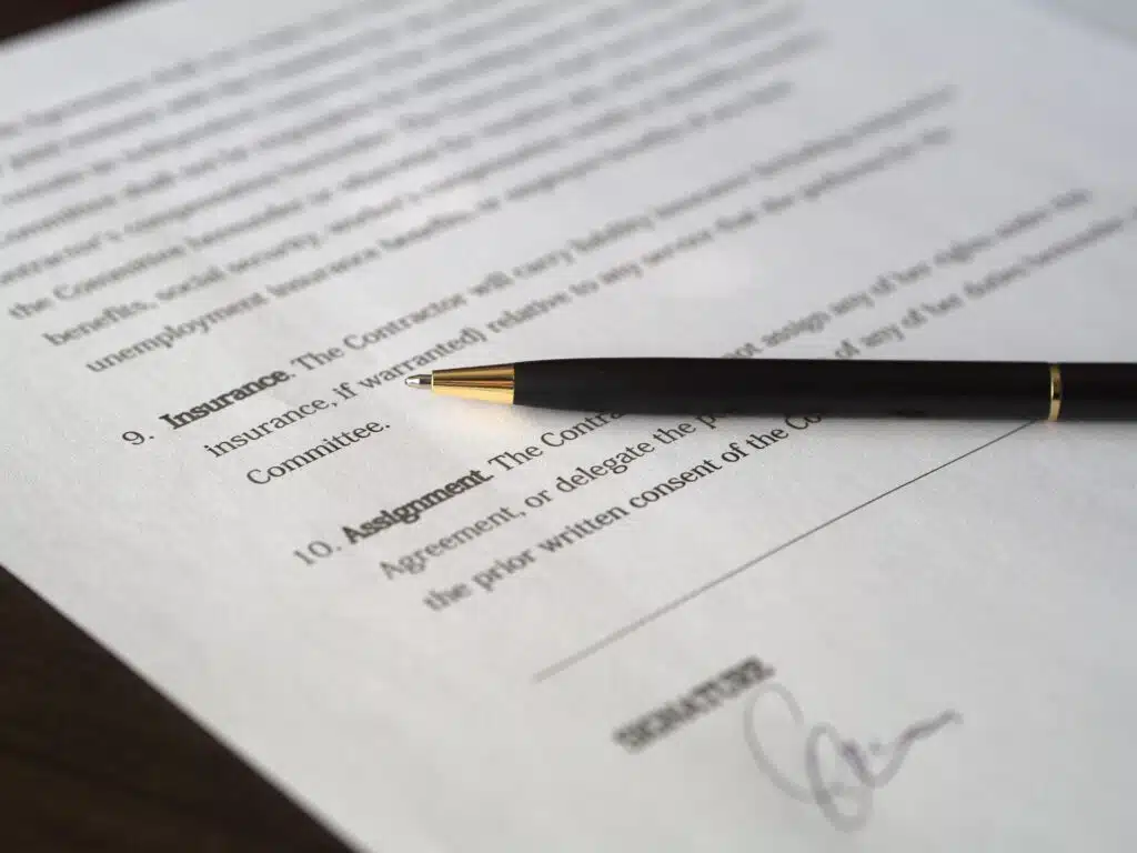 A black pen rests on a document with numbered sections titled "Insurance" and "Assignment." There's a signature near the bottom, indicating the document's potential legal or formal nature.